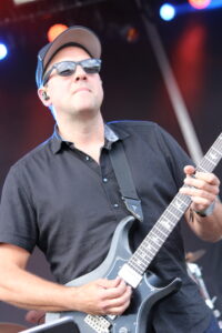Male guitarist wearing sunglasses and a cap, playing a gray electric guitar on a concert stage.