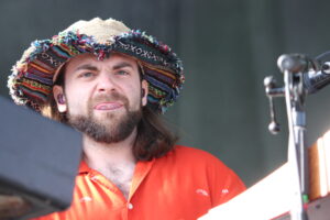 Close-up of a man with a beard wearing a colorful woven sombrero and an orange shirt, standing at a microphone on stage.