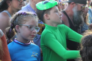 Young girl with glitter in braided purple hair and blue shirt among a crowded outdoor event crowd.