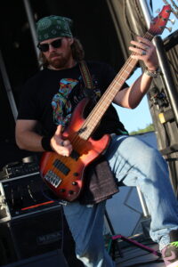 Bass guitarist performing on stage, wearing a green bandana, sunglasses, and a black graphic tee, playing a sunburst bass guitar.