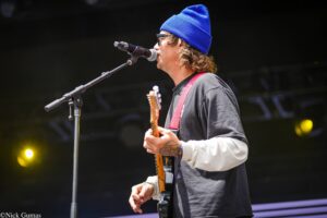 Guitarist in a blue beanie performs on stage, singing into a microphone while playing an orange electric guitar with a red strap.