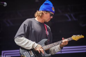 Guitarist wearing a blue beanie and sunglasses, playing a black electric guitar on a dim stage.