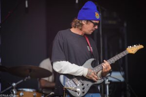 Guitarist wearing a blue beanie and sunglasses plays a black electric guitar on stage, with a drum set visible in the background on the left.