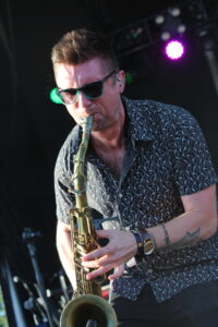 Male saxophonist wearing teal sunglasses and a patterned short-sleeve shirt, playing a saxophone on a stage with purple spotlight in the background.
