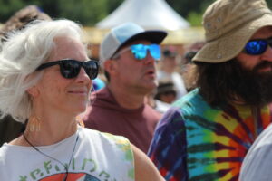 Older woman with white hair wearing black sunglasses at an outdoor crowd event, smiling slightly.