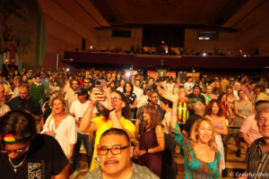 Crowd of people at a lively indoor concert, facing the stage with some raising phones and drinks in hand, warm lighting and a balcony in the background.