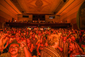 Large indoor crowd at a concert under orange/red lighting, people smiling, some with hands raised, balcony area visible in the background.