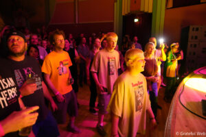 Crowd of concertgoers facing the stage under pink and orange lighting in a club venue, some holding drinks and phones.