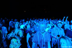 Crowd at a concert bathed in blue lighting, people smiling and raising their hands toward the stage.