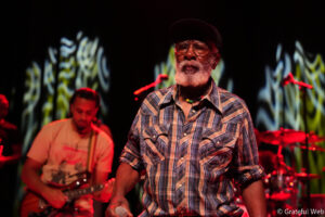 Older man with glasses and a beard sings into a microphone on stage, wearing a plaid shirt and cap (live performance).