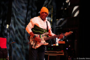 Male guitarist on stage wearing an orange beanie and white hoodie, playing a sticker-covered electric guitar during a live performance