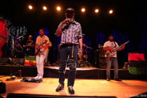 Lead singer in a plaid shirt and ripped jeans sings into a microphone on a dimly lit stage, with band members playing instruments in the background.