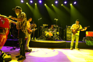 Band performs on a brightly lit stage; guitarist center-left, bassist right, drummer behind them under yellow/purple lights; foreground foot and cables visible.