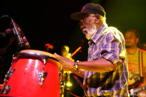 Older man with glasses and a white beard plays red conga drums on stage under warm yellow lights during a live performance.
