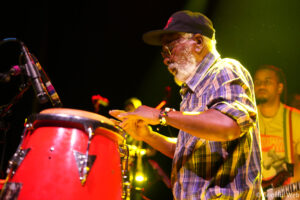 Male percussionist with a white beard wearing a dark cap and plaid shirt, playing red conga drums on a brightly lit stage, microphone in foreground.