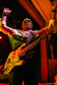 Musician on stage with arms raised, wearing an orange beanie and colorful hoodie, playing a sticker-covered electric guitar under warm stage lights
