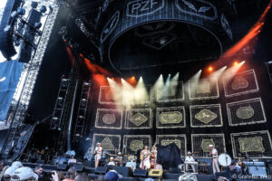 Live concert on a large outdoor stage with musicians performing and colorful stage lights, audience visible in front.