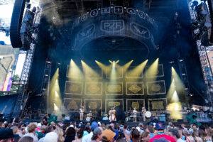 Outdoor concert stage at a festival with yellow spotlights and a cheering crowd in front of a large, illuminated backdrop.