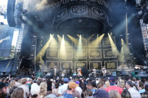 Outdoor music festival stage with bright spotlights and a full crowd watching a live band beneath a large rigging structure.