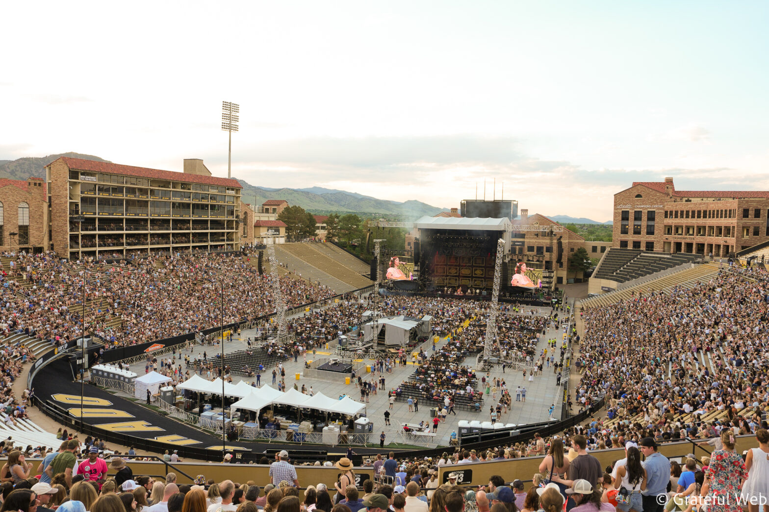 Overflowing outdoor stadium filled with concertgoers facing a large stage with screens and lights.
