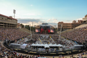 Large outdoor stadium concert with a central stage, big screens, and a packed audience under a blue sky.