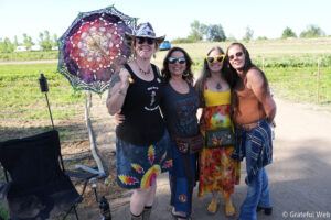 Four smiling women posing outdoors in a sunny rural field; one holds a colorful lace parasol.