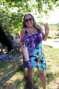 Smiling woman in a colorful tie-dye dress making a peace sign outdoors, pink tumbler in hand, sunglasses on, with trees in the background.