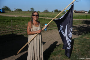 Woman in a beige dress wearing sunglasses holds a long wooden flagpole with a dark flag in a sunlit field.