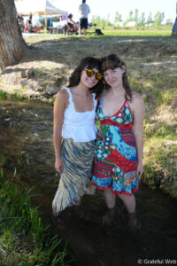 Two women stand in a shallow creek, smiling for a photo, wearing colorful summer dresses with sunglasses on a sunny day near a park.