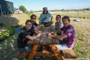 Five friends sit around a wooden picnic table outdoors on a sunny day, smiling at camera in a grassy field with tents in the backgrounde