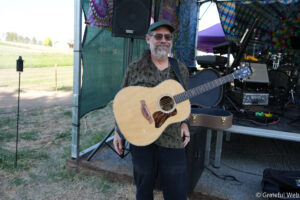 Man with gray beard and glasses holding an acoustic guitar on an outdoor stage with colorful festival backdrop behind him, smiling to the camera.