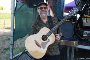 Bearded guitarist wearing a green cap and patterned shirt playing an acoustic guitar on an outdoor stage with equipment and a colorful backdrop behind him.