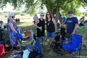Smiling group of adults standing under a large tree at a sunny outdoor picnic, with folding chairs and coolers nearby.