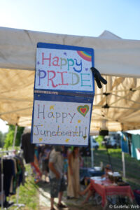 Colorful 'Happy PRIDE' sign with a rainbow accent hanging from a tent at an outdoor market.