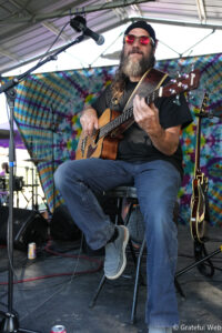 Musician with a beard and red sunglasses playing an acoustic guitar on a stage with a tie-dye backdrop.