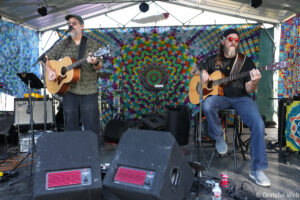 Two bearded musicians on stage playing acoustic guitars, one singing into a microphone, colorful tie-dye backdrop behind them.