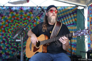 Bearded man in a black beanie and red sunglasses playing an acoustic guitar on a colorful tie-dye stage backdrop with a microphone in front.