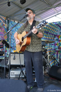 Male musician with gray beard and cap playing an acoustic guitar on a colorful stage backdrop.