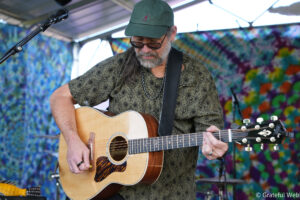 Male guitarist with sunglasses and a green cap plays an acoustic guitar on a colorful stage backdrop.