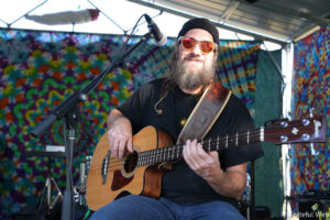 Bearded guitarist wearing a black beanie and orange sunglasses, playing an acoustic guitar on a tie-dye stage backdrop.