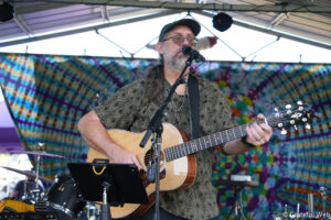 Male guitarist with long hair and beard singing into a microphone while playing an acoustic guitar on a colorful tie-dye stage backdrop.