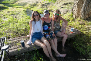 Three women sit on a weathered log beside a small creek, smiling at the camera on a sunny day.
