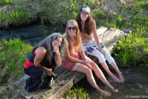 Three women sit on a weathered log beside a narrow creek, smiling at the camera in a sunny grassy area.