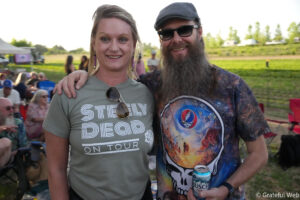 Two smiling adults stand arm in arm at an outdoor event; woman on left wears a green shirt and sunglasses, man on right has a cap, sunglasses, and a colorful graphic shirt while holding a Busch beer can in a sunny park setting.