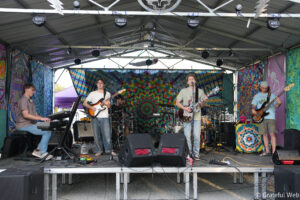 A four-piece band performing on an outdoor stage with guitars, keyboard, drums, and a colorful tie-dye backdrop behind them.
