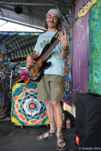 Bassist performing on a colorful tie-dye stage backdrop, wearing a bandana and light blue shirt.
