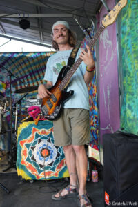 Bass guitarist smiling while playing on a colorful tie-dye stage backdrop, wearing a light blue t-shirt and a bandana.