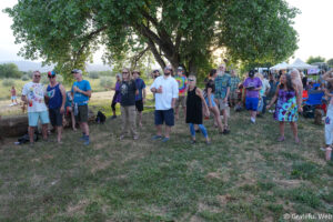 Group of adults standing and watching something outdoors under a large tree at a festival or gathering, with tents in the background