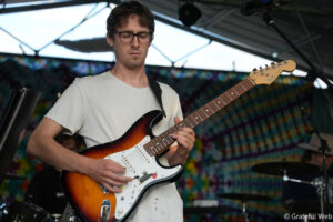 Guitarist with glasses and white shirt playing a sunburst Fender Stratocaster on a stage with colorful backdrop behind him.