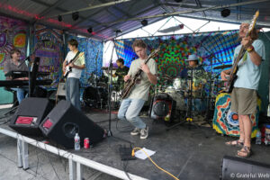 A live band performing on a colorful tie-dye stage with guitars, drums, and keyboards under a metal canopy.